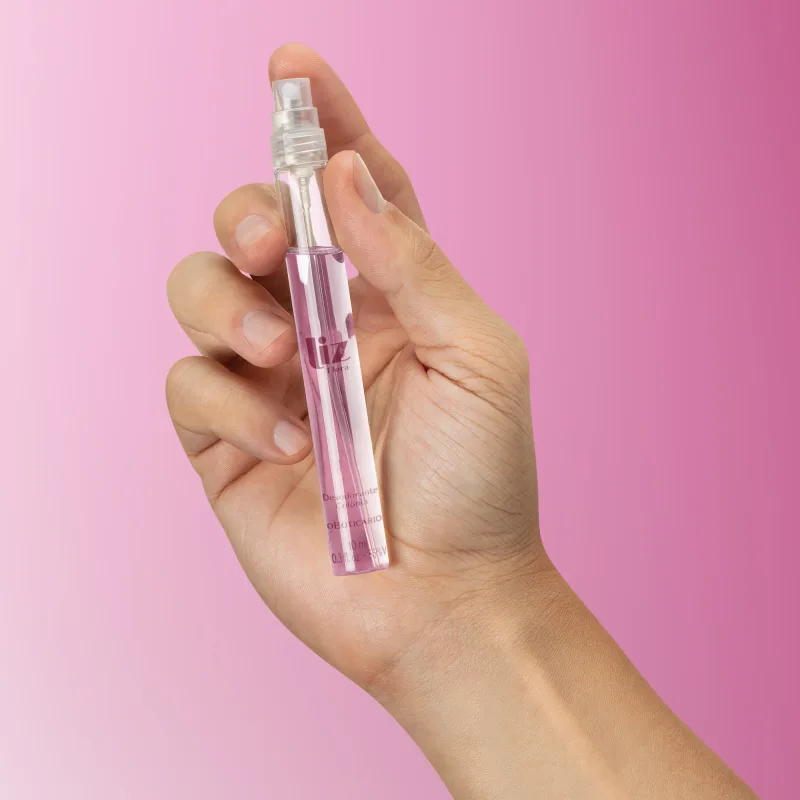 Mão feminina segurando um frasco cilíndrico transparente com líquido claro, tampa preta e letras na cor vinho contendo 10ml de desodorante colônia Liz Flora em fundo em tons de roxo.