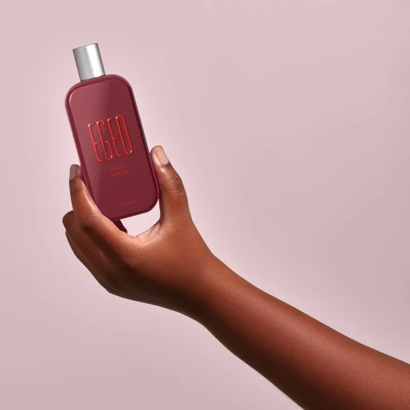 Frasco vermelho de desodorante colônia Egeo Choc High sendo segurado por mão feminina de pele negra contra fundo rosa claro.