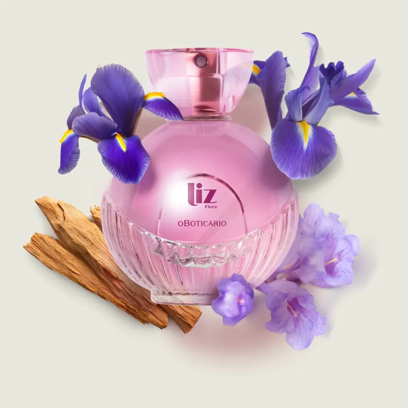Vidro arredondado com líquido rosa, letras vinho, borrifador dourado e tampa transparente contendo desodorante colônia Liz Flora rodeado por flores de íris violetas e pedaços de madeira em superfície clara.
