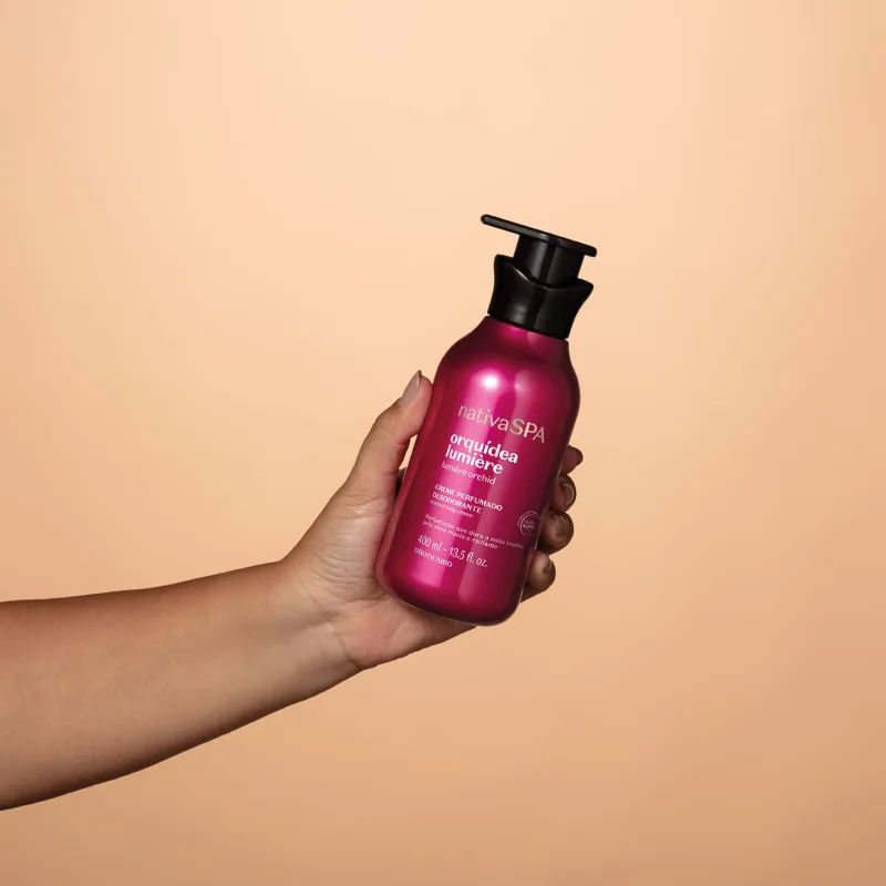 Embalagem pump rosa brilhante com tampa preta de item de corpo e banho da marca Nativa SPA Orquídea Lumiere sendo segurada por mão feminina em fundo laranja claro.