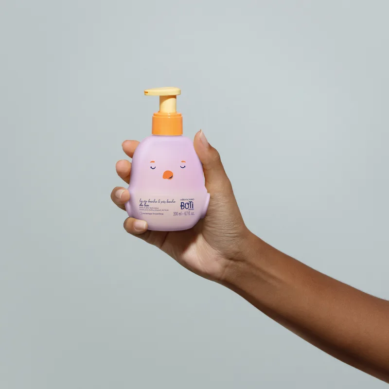Mão feminina segurando um frasco da loção para banho e pós banho Boti Baby Lua em forma de pintinho na cor lilás e bomba dosadora laranja em fundo lilás.