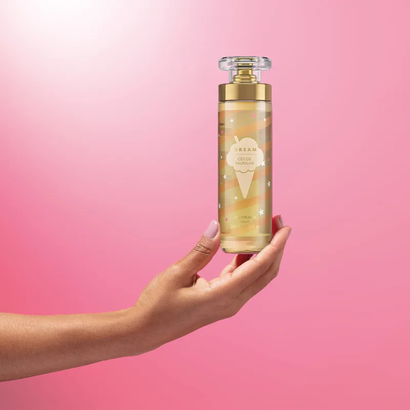 Imagem de uma mão segurando um frasco de body splash Dream Céu de Baunilha. A embalagem conta com tampa transparente, válvula spray dourada e rotulo em tons de dourado. O cenário traz um fundo em tons de lilás e branco.