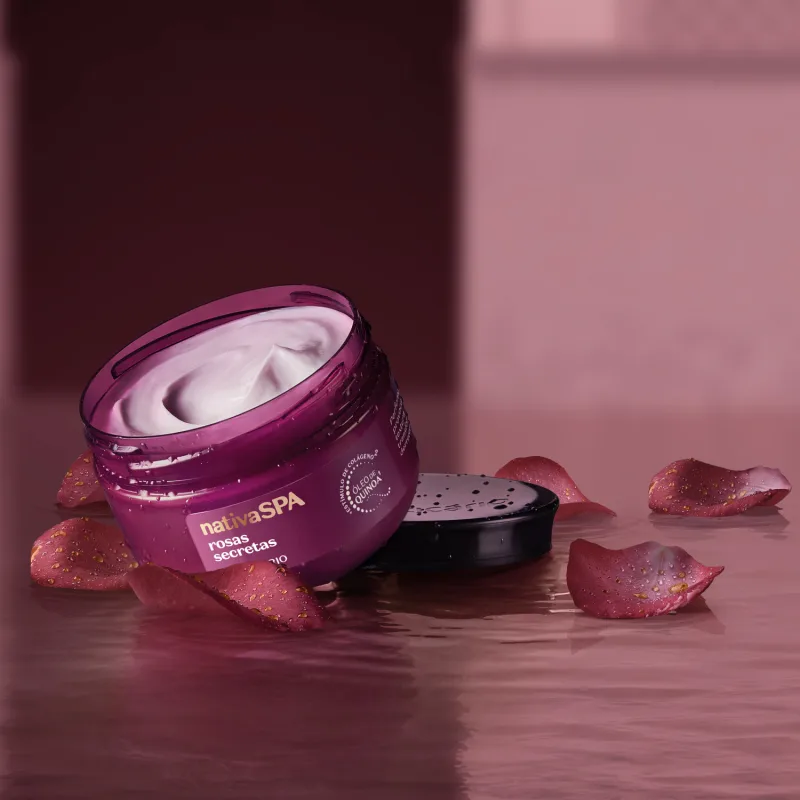 Ambiente de spa com um pote de óleo em creme Nativa SPA Rosas Secretas. Ele está sobre pétalas de rosa espalhadas sobre uma superfície úmida, em um cenário monocromático de tons de roxo.