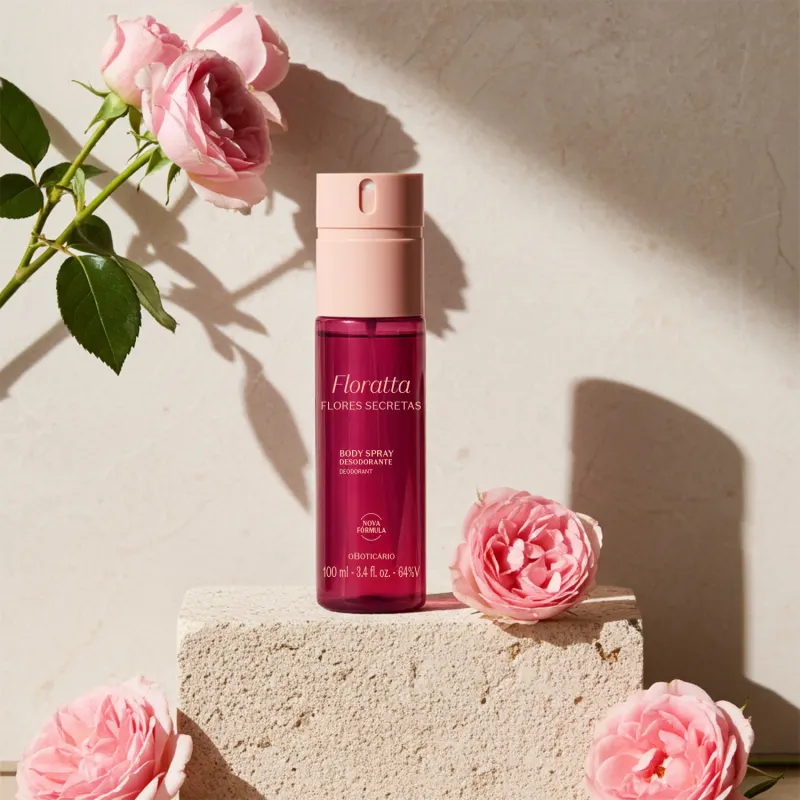Imagem de embalagem de body spray da marca Floratta Flores SEctas em frasco vinho com tampa rosa,, posicionada sobre superfície de concreto, com rosas cor de rosa ao redor, em ambiente iluminado com sombras suaves.
