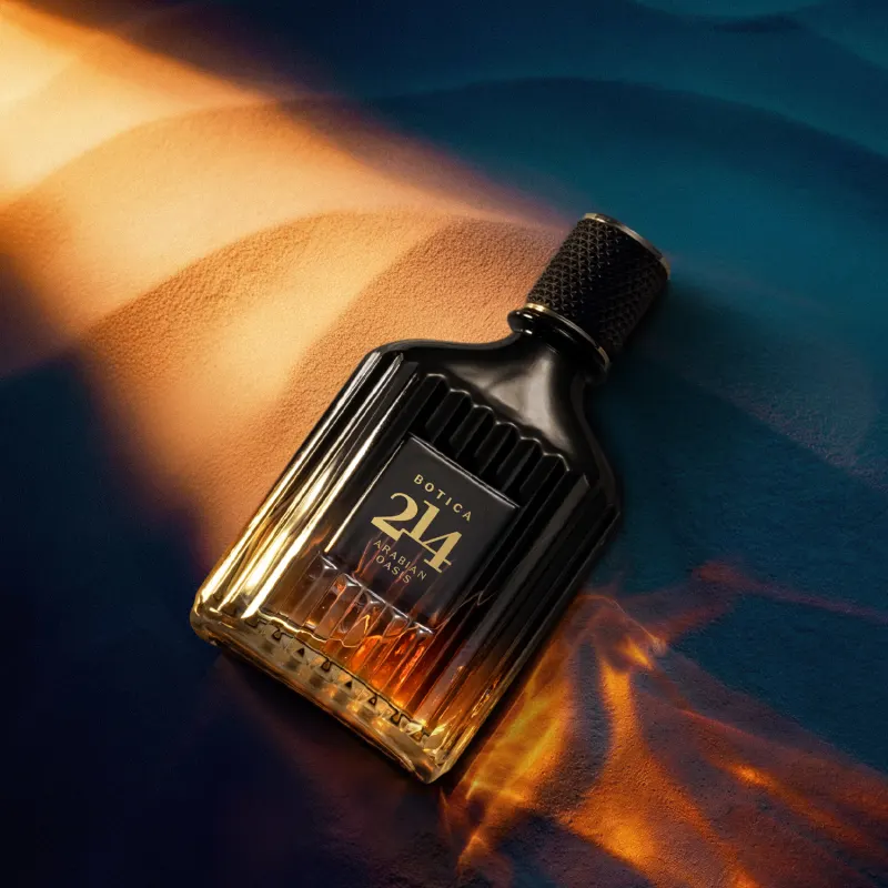 Frasco de item de perfumaria da marca Botica 214 Arabian Oasis em primeiro plano, sobre superfície de areia ondulada desfocada e iluminação cenográfica dramática.