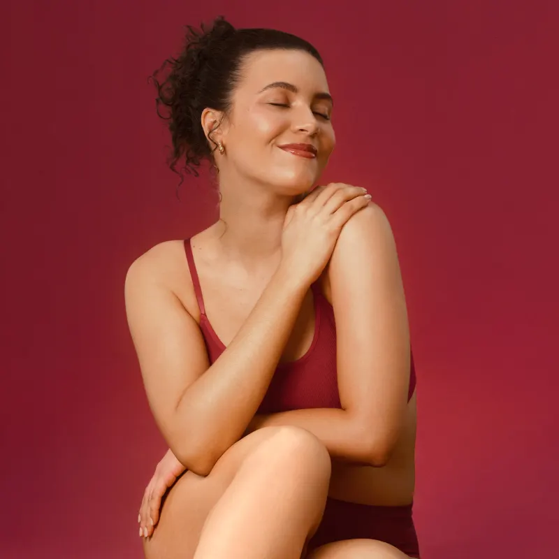 Imagem de mulher de cabelos cacheados usando top vermelho, sentada com os braços cruzados em um fundo vermelho. Ela espalha um pouco de loção hidratante corporal no ombro. Está sorrindo e de olhos fechados.