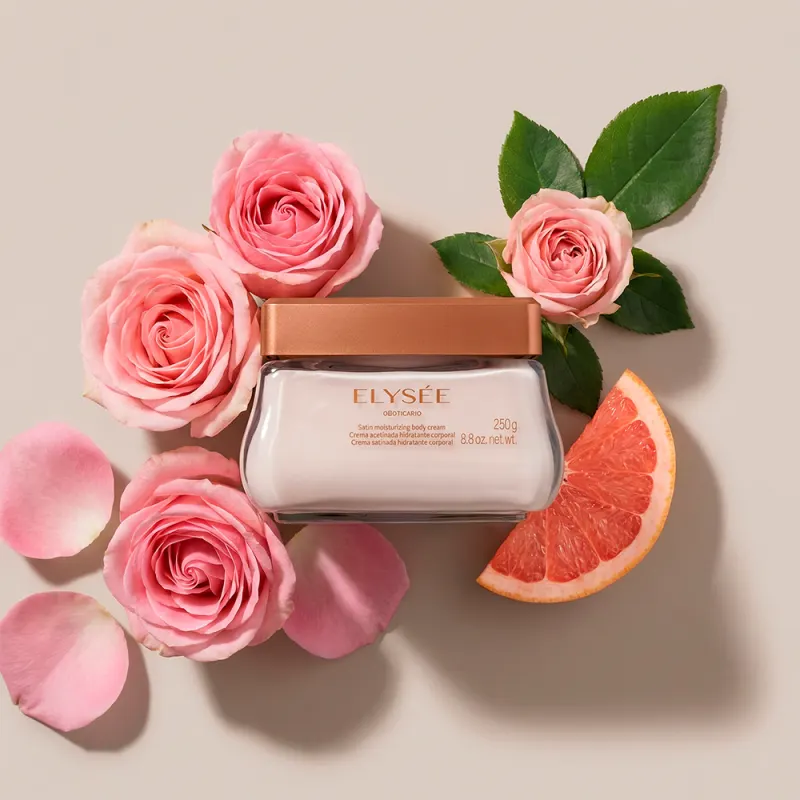 Imagem do pote de creme corporal da marca Elysée em primeiro plano, rodeado por rosas cor de rosa e fatia de toranja sobre fundo bege claro. O ambiente sugere um contexto de feminilidade, delicadeza e frescor.
