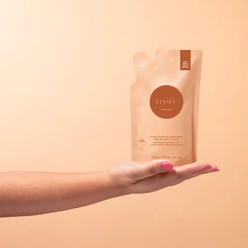Imagem de uma mão segurando um refil de creme hidratante desodorante corporal da marca Elysée, em embalagem de sachê com predomínio de tons de laranja e marrom, contendo informações sobre a fórmula e o volume do produto. O fundo é em tons de rosa.
