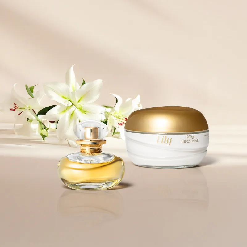Imagem de um frasco dourado de Eau de Parfum Lily e um pote branco com tampa dourada de creme corporal da mesma marca em fundo e superfície claras e lírios brancos ao lado.