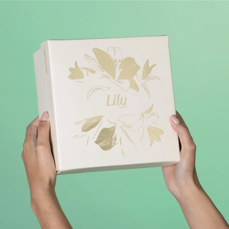 Imagem de uma caixa branca com ilustração dourada de flores, sobre fundo verde claro, segura por mãos femininas.