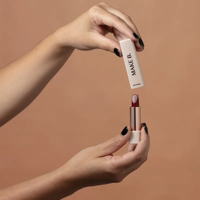Uma mão feminina segura a tampa branca do batom cremoso Make B. na cor vermelha, enquanto a outra mão segura o produto produto em tom dourado em fundo bege.