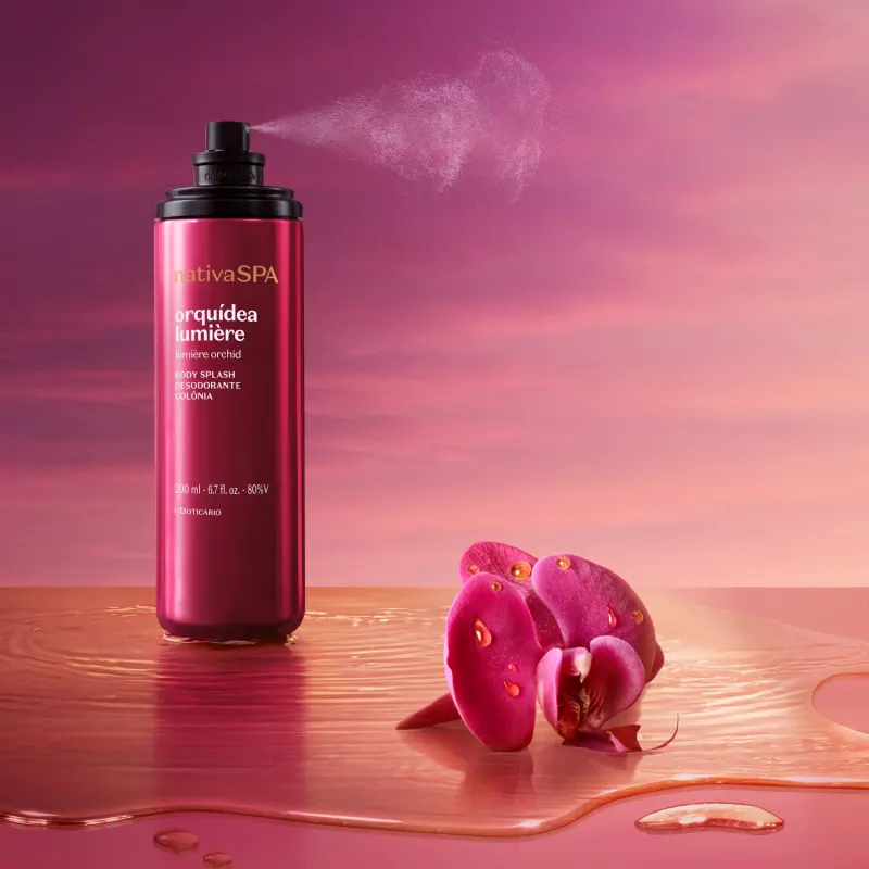 Frasco de item de perfumaria da marca Nativa SPA Orquídea Lumiere aberto e da válvula spray vemos sair uma bruma do produto. O frasco na cor rosa está sobre superfície rosa molhada e ao lado, uma orquídea rosa contra um céu rosa e laranja.