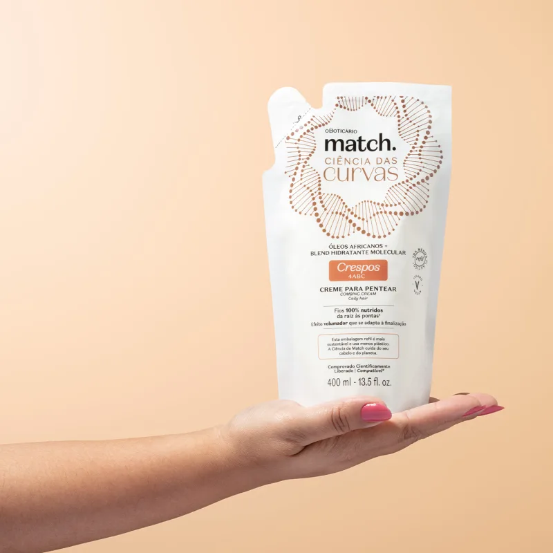 Imagem de uma mão segurando uma embalagem refil branca de creme para pentear da marca Match, com design de círculos e linhas em marrom, informando "Ciência das Curvas" e "Crespos 4ABC" no rótulo. O fundo é rosa bem claro.