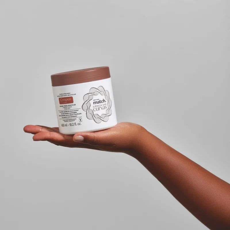 Imagem de uma mão segurando um pote branco com tampa marrom contendo creme para pentear da linha Match. Ciência das Curvas Crespos em fundo cinza.