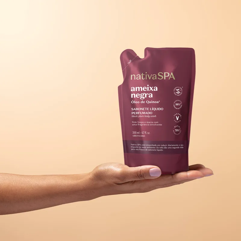 Imagem de uma mão segurando uma embalagem refil de sabonete líquido perfumado da marca Nativa SPA, na cor vinho, com rótulo destacando o nome "Ameixa Negra". O fundo é em tons de branco e rosa.