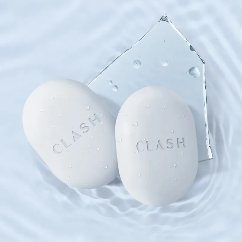 Dois sabonetes brancos com a marca "Clash" em relevo, flutuando sobre uma superfície aquática com gotas d'água, em uma superfície clara e molhada