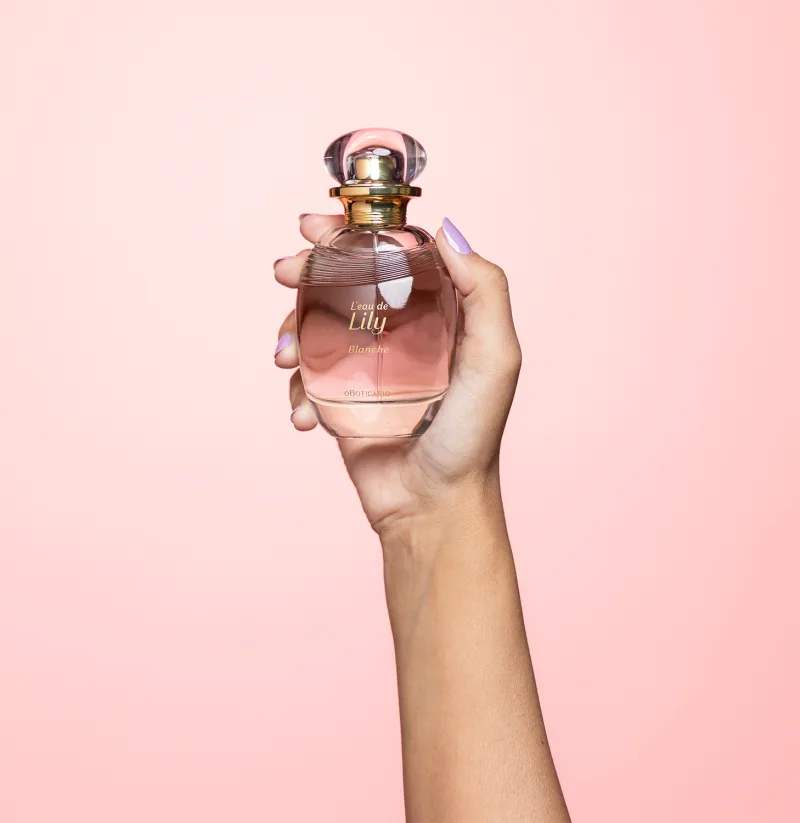 A cor predominante do frasco é um tom rosado, que transmite uma sensação delicada e feminina. A fragrância é segurada por uma mão feminina, com unhas pintadas em uma cor clara, indicando se tratar do desodorante colônia L'eau de Lily Blanche.