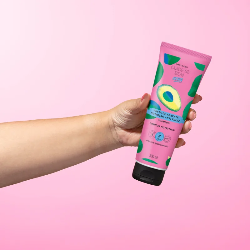 Imagem de um tubo roxo com ilustrações de abacate de shampoo da marca Cuide-se Bem Feira Pasta de Abacate, seguro por mão em fundo rosa.