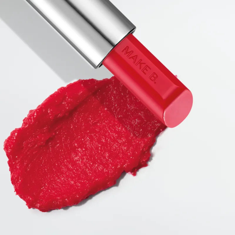 O balm labial vermelho da marca Make B. Lip Chronology Color Repair possui uma embalagem metálica prateada e produto vermelho brilhante. Há uma mancha do item sobre fundo branco, indicando a textura e a intensidade do produto.