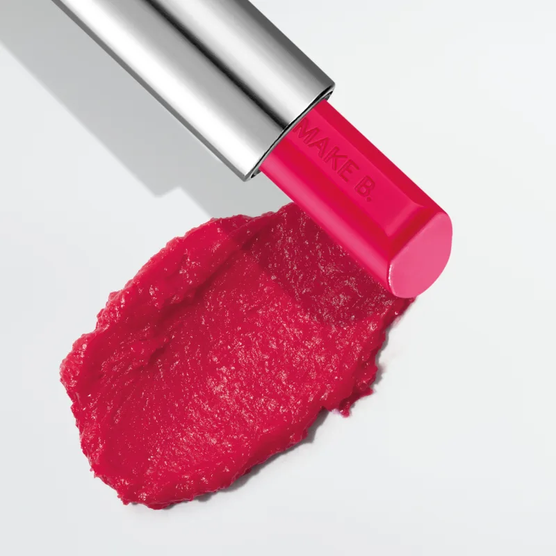 O balm labial rosa da marca Make B. Lip Chronology Color Repair possui uma embalagem metálica prateada e produto cor de rosa brilhante. Há uma mancha do item sobre fundo branco, indicando a textura e a intensidade do produto.
