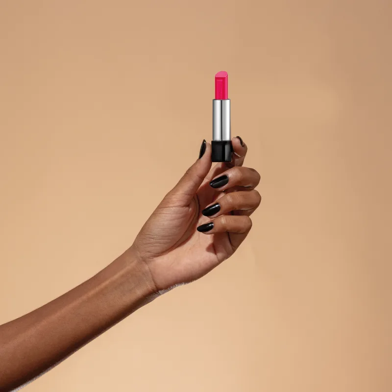 Uma mão feminina segura um balm labial rosa da marca Make B. Lip Chronology Color Repair. O produto está centralizado contra um fundo laranja claro.
