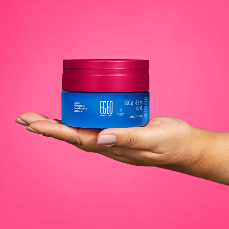 Imagem de uma mão segurando um pote azul com tampa rosa contendo 250g de creme hidratante desodorante corporal da marca Egeo Dolce Illusion. O fundo da imagem é na cor rosa liso.