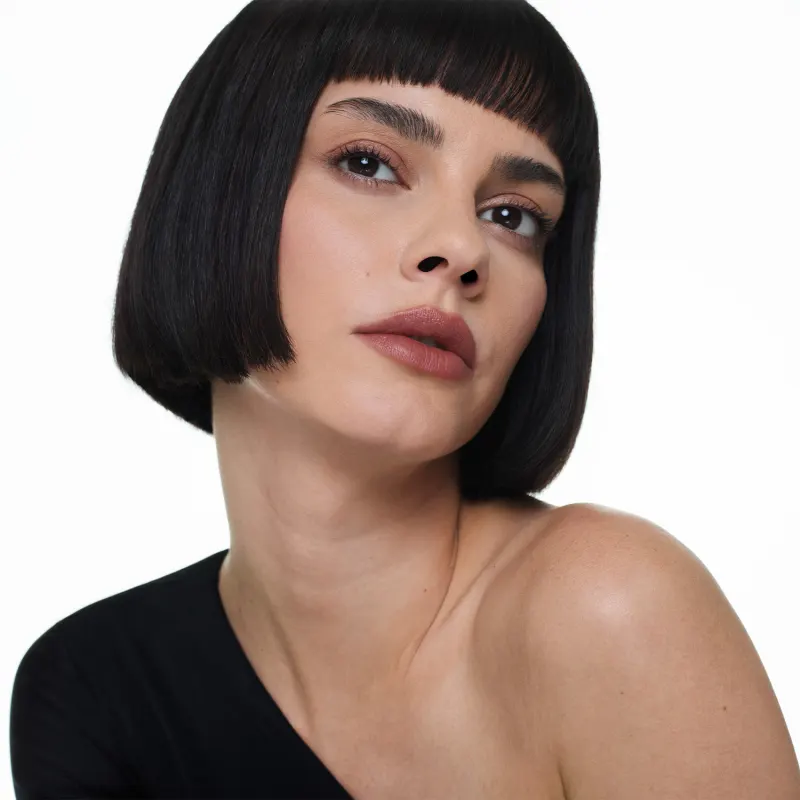 Mulher branca de cabelo preto curto e franja, com expressão séria, usando roupa preta, em fundo branco om olhar sério e lábios levemente entreabertos pintados com batom marrom claro.