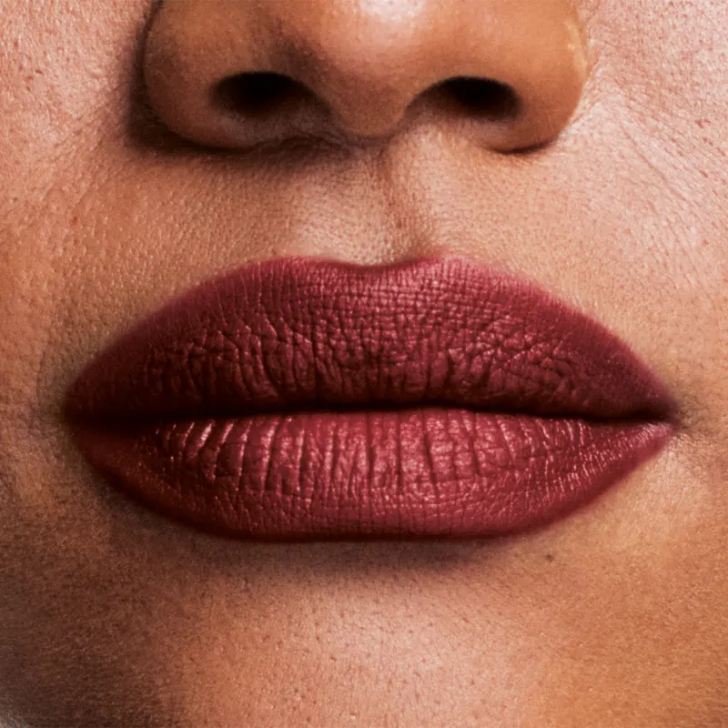 Boca feminina de mulher branca em close mostrando queixo e um pedaço do nariz, com lábios fechados com batom avermelhado sobre eles.