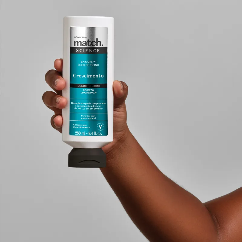 Embalagem de condicionador na cor branca com rotulo azul da linha Match Science Crescimento. O produto está sendo segurado de frente por mão feminina negra em fundo cinza claro.