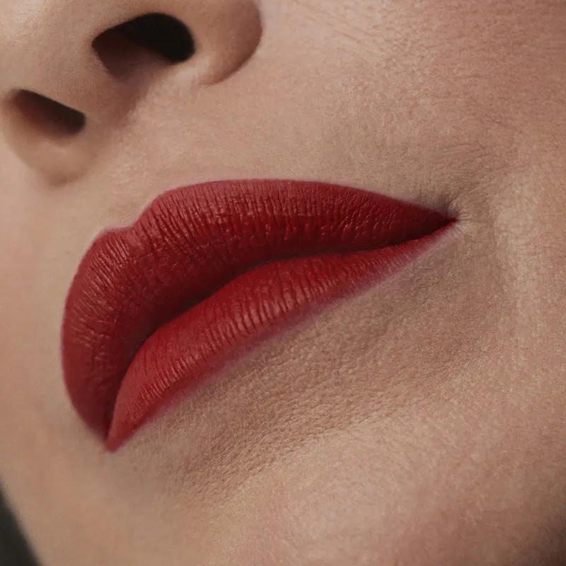 Imagem de um rosto feminino em close-up, evidenciando os lábios com o batom líquido Make B. em tom vermelho bem forte. A boca está fechada com um semblante sério.