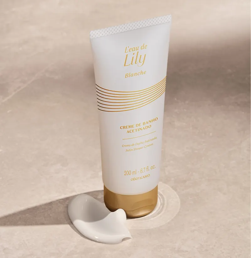 Imagem de um frasco branco com tampa e rótulo dourados de creme de banho acetinado da linha L'eau de Lily Blanche em fundo bege com sombras e pequena quantidade do produto ao lado da embalagem.