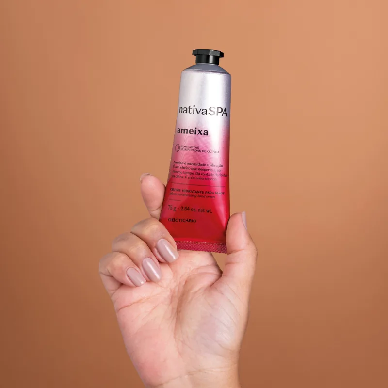 Imagem de uma mão feminina com unhas longas segurando um tubo de creme hidratante para as mãos Nativa SPA Ameixa. A bisnaga é na cor prata e rosa, possui tampa preta e informações sobre o produto estão impressas em letras pretas.. O fundo é bege.