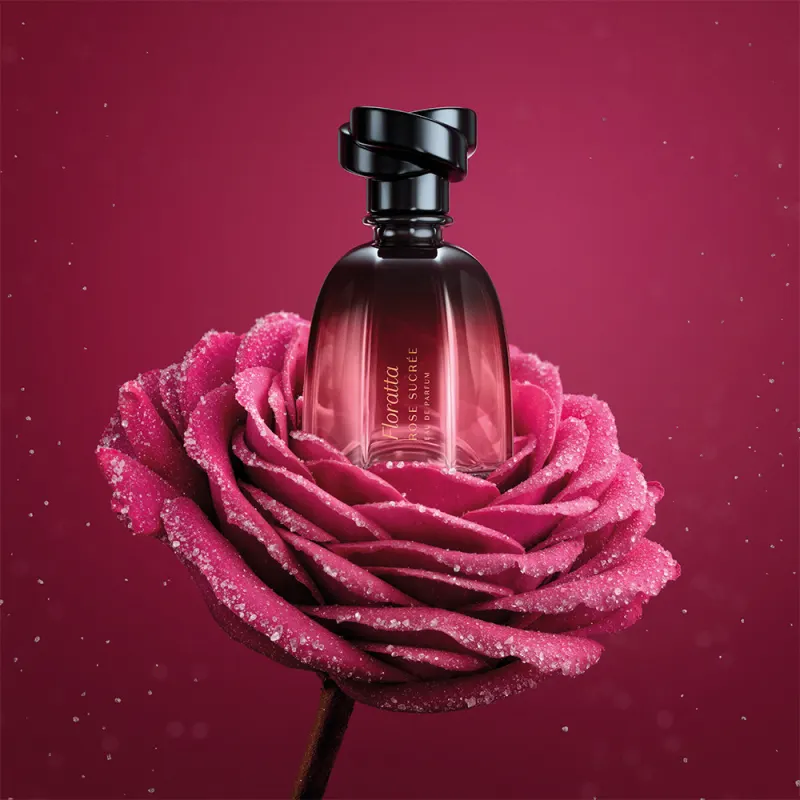 Frasco de item de perfumaria Floratta Rose Sucrée em cima de uma rosa sobre fundo vermelho-escuro. As pétalas são rosa brilhantes e possuem gotículas de orvalho.