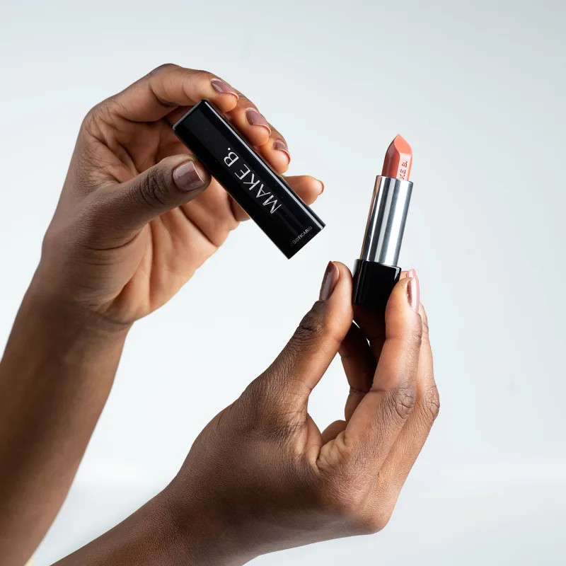 Mãos femininas segurando a tampa preta de um batom da marca Make B. na mão direita e a base do batom com a bala aparente na mão esquerda, em um ambiente minimalista e claro.
