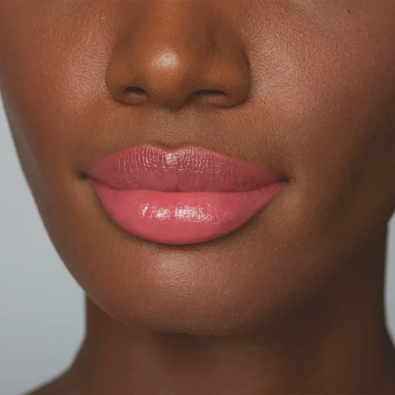 A imagem mostra parte de um rosto feminino em primeiro plano, dando destaque aos lábios, mostrando como o batom rosa da /Make B. fica na pele negra. O fundo é cinza e liso.
