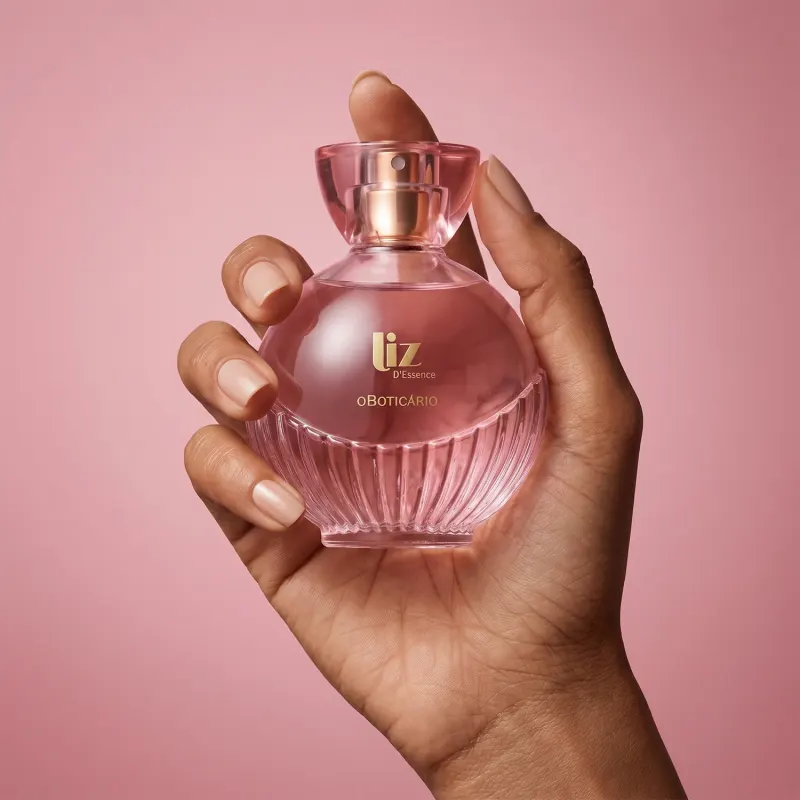 Imagem de um frasco de perfume rosa da marca Liz d'Essence, em uma mão feminina, em um fundo rosa sólido, sugerindo um ambiente de beleza e cosmética.