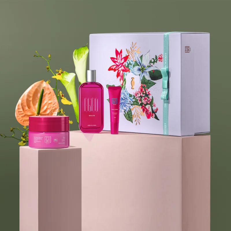 Imagem do kit presente Egeo Dolce com creme hidratante corporal, desodorante colônia com tampa prateada, bisnaga de gloss labial e caixa branca com ilustração floral.  O kit está em cima de pilastras rosa, com flores amarelas e branca e fundo cinza.