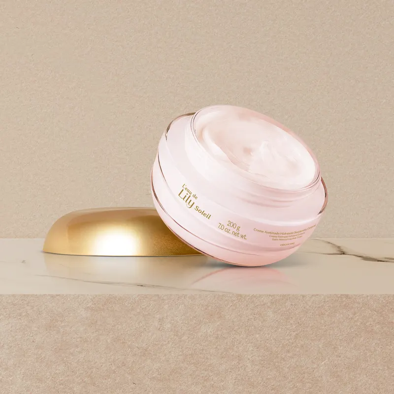 Imagem do pote transparente de creme acetinado corporal L'eau de Lily Soleil aberto sobre superfície de mármore bege, com a tampa dourada ao lado.