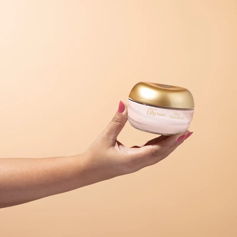 Imagem de uma mão feminina segurando um pote de vidro transparente com conteúdo branco e tampa dourada contendo creme acetinado corporal da marca L'eau de Lily Soleil em fundo bege vibrante.