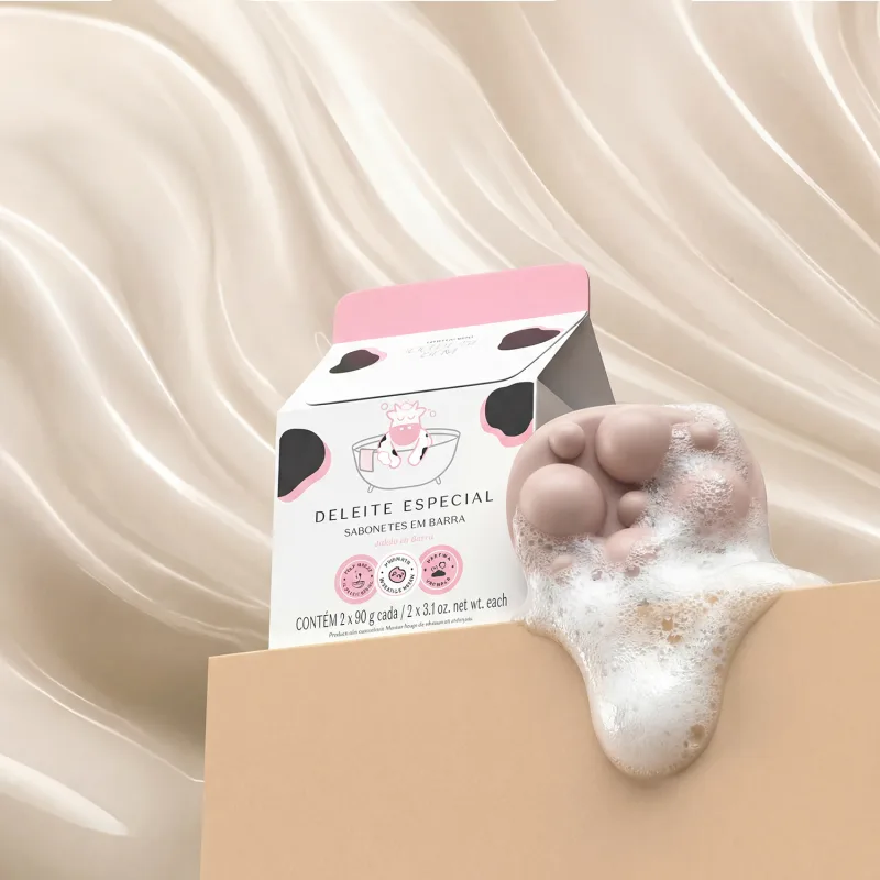 A imagem mostra a caixa branca estampada do sabonete em barra Cuide-se Bem Deleite e dois sabonetes em uma superfície rosa. O produto está cheio de espuma branca que escorre. O fundo é rosa com texturas.