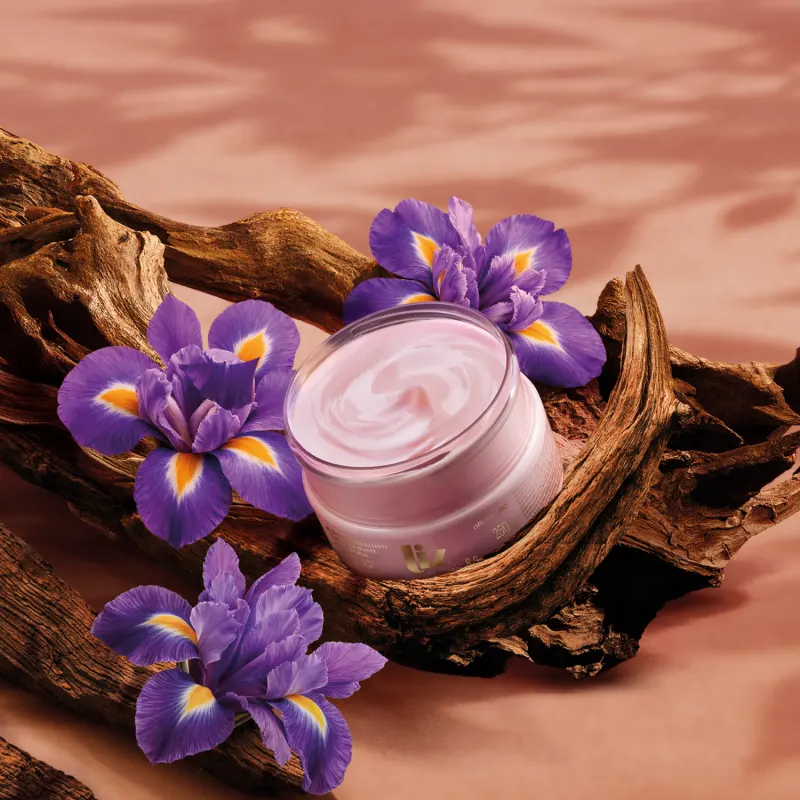 Recipiente de item de perfumaria rosa em meio a flores de íris roxas sobre troncos de madeira em cenário com céu alaranjado.