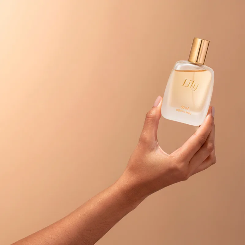 Imagem de um frasco de vidro da fragrância Lily em embalagem transparente com tampa e letras douradas, sendo segurado por uma mão feminina com fundo bege.