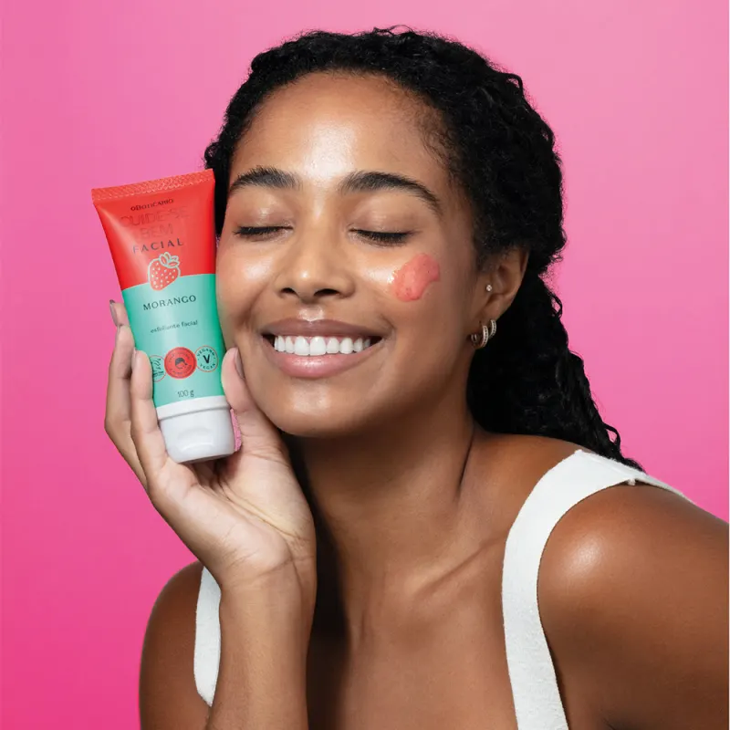 Imagem de uma mulher segurando a bisnaga de esfoliante facial Cuide-se Bem Morango. A embalagem é vermelha e azul-turquesa e está bem próxima ao rosto da mulher que sorri e tem os olhos fechados. seu cabelo é preto e ela usa um top branco. O fundo é rosa.