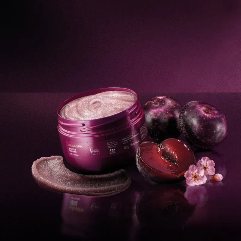 Pote de creme cosmético rosa em superfície escura, com flores e frutos roxos ao redor, em ambiente de penumbra com tons de roxo e preto.