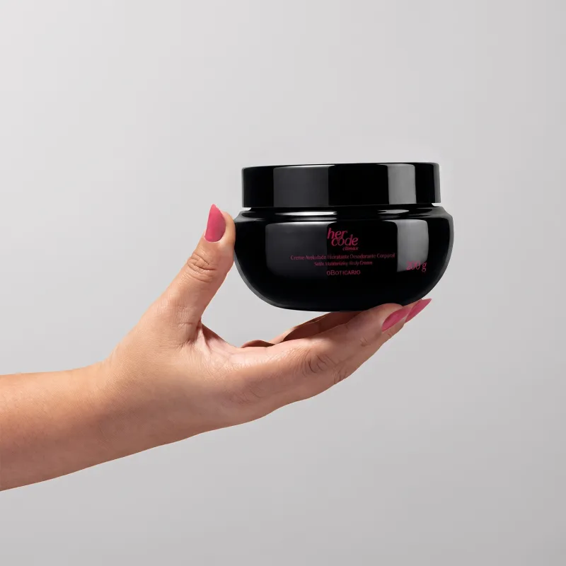 Recipiente preto de item de corpo e banho da marca Her Code, seguro por mão feminina com unhas pintadas de rosa, sobre fundo branco liso.