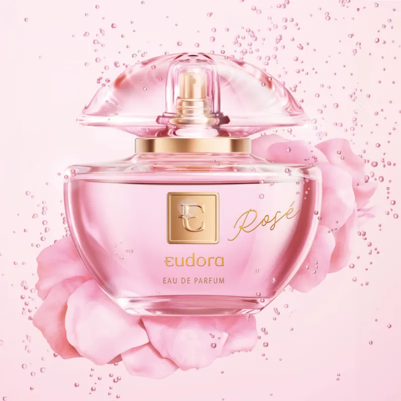 A imagem mostra o frasco de vidro transparente em tons de rosa com o nome "Rose" escrito em letras cursivas e o logotipo da Eudora. O frasco está cercado por gotículas de espumante e pétalas de rosa espalhadas, criando uma ambientação delicada e feminina.