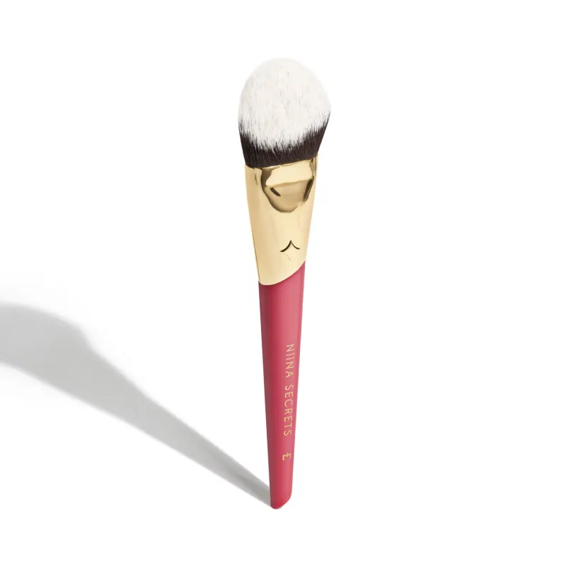 A imagem mostra um pincel de maquiagem, visto de um ângulo um pouco mais elevado, sem ser exatamente de frente. Com cabo vermelho e dourado, e cerdas pretas com ponta branca. Na parte vermelha, logo da marca e linha do produto escrito em dourado.
