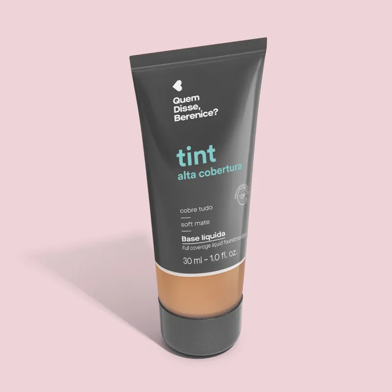 A imagem mostra a Base Tint Alta Cobertura da Quem Disse, Berenice? Embalagem cinza escura com parte inferior transparente para mostrar a cor do produto sobre fundo rosa.