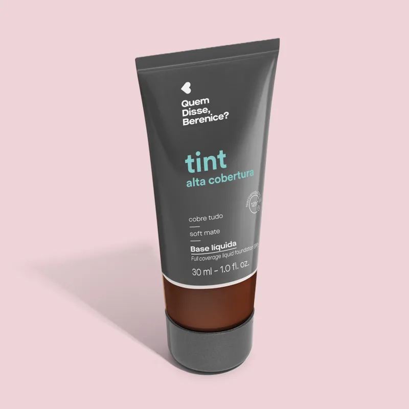 A imagem mostra a Base Tint Alta Cobertura da Quem Disse, Berenice? Embalagem preta com parte inferior transparente para mostrar a cor do produto sobre fundo rosa.