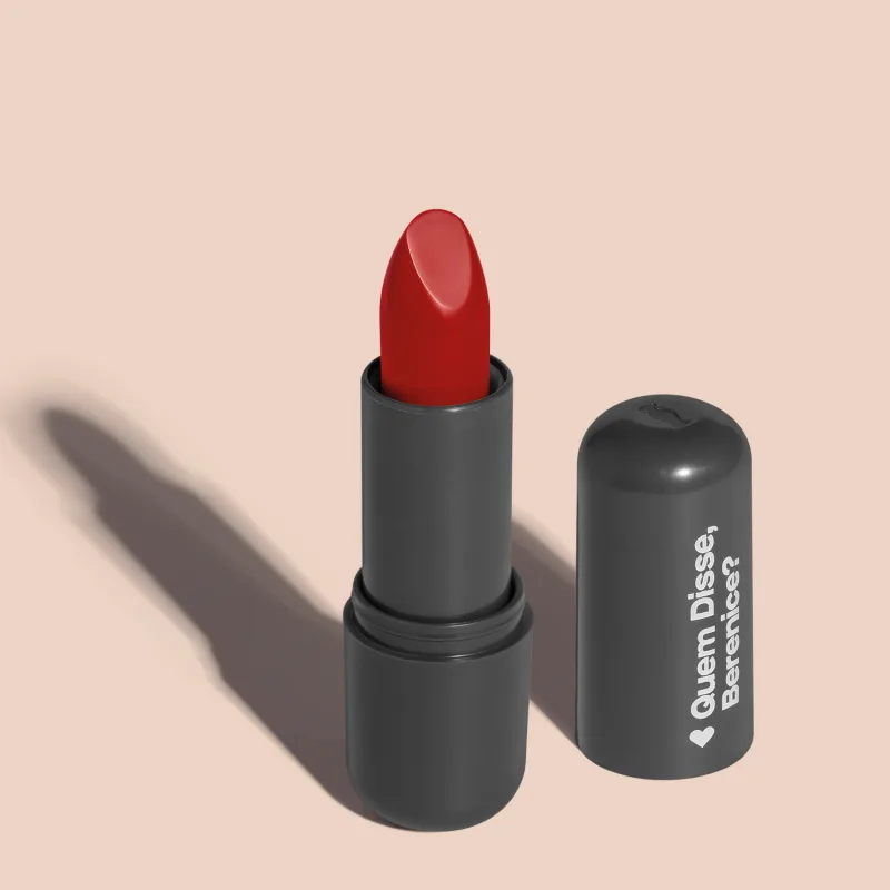 Batom de cor vermelho vibrante em embalagem preta com logotipo da marca Quem Disse, Berenice? em destaque. O batom está aberto sobre superfície rosa com iluminação que cria uma longa sombra projetada atrás da embalagem.